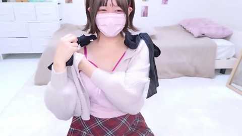 Video of an Asian woman in a white mask, pink blouse, and red plaid skirt, adjusting a gray cardigan, in a modern bedroom with white furniture, fluffy pillows, and a bed.