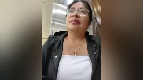 Video of a smiling Asian woman with glasses, wearing a black blazer over a white top, standing in front of a mirrored wall.