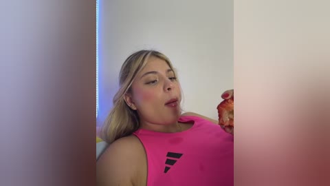Video of a blonde woman with fair skin, wearing a pink Adidas sports bra, drinking a glass of orange juice. She has a flushed face and looks relaxed.