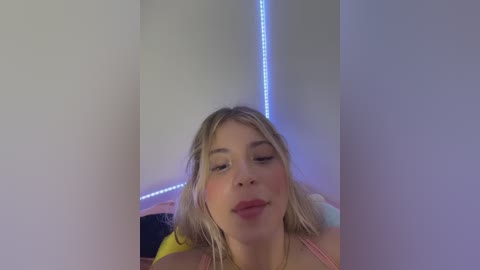 Video of a young woman with fair skin, blonde hair, and light makeup, wearing a pink top, sitting on a bed with neon blue lights in the background.