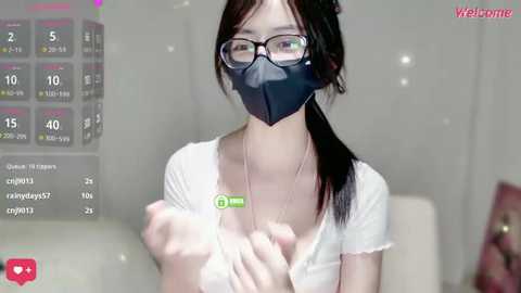 A video of an Asian woman with long black hair, wearing a white shirt, black face mask, and glasses, holding a green box in a modern room with a calendar overlay.
