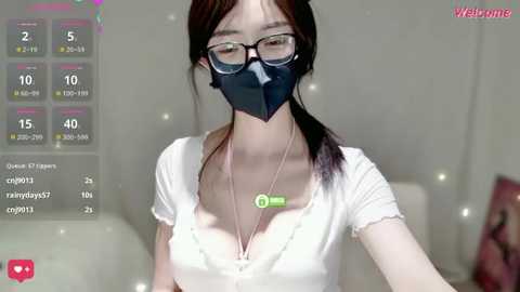 A video of an East Asian woman with light skin, long black hair, and glasses, wearing a black mask, white top, and necklace, in a bedroom setting.
