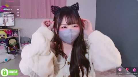 Video of an Asian woman with long black hair, wearing a white furry jacket, black cat ears, and a blue surgical mask, adjusting her hair in a cozy room with shelves and stuffed toys.