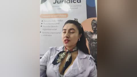 Video of a woman with medium skin tone and dark hair, wearing a white shirt with a black floral pattern, leaning against a poster advertising a \"Juriada\" event. She has a seductive expression.