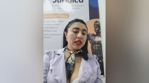 Video of a young woman with medium skin tone, straight black hair, wearing a light purple blouse, unbuttoned to reveal a black bra, and a patterned scarf. Background includes a medical brochure with text in Spanish.