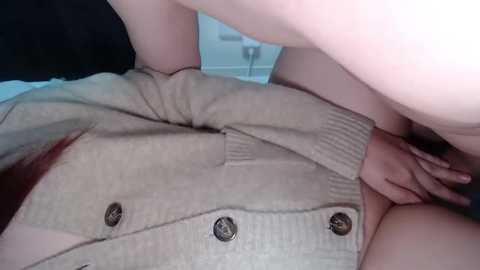 Video of a light-skinned person in a beige cardigan with black buttons, lying on a bed with white sheets, legs spread, and genitals visible.