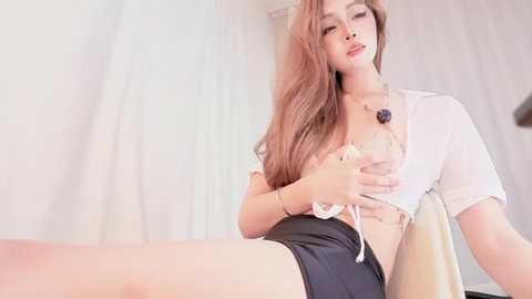 Video of an attractive Asian woman with long, wavy, light brown hair, wearing a white crop top and black shorts, seated on a white chair, touching her breasts.