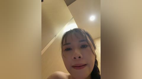 A low-angle, close-up video of a young Asian woman with long black hair and fair skin, smiling slightly, captured indoors with beige walls and a recessed ceiling light.