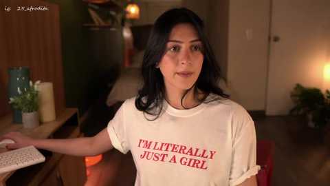 Video of a Latina woman with long black hair, medium skin tone, wearing a white t-shirt with \"I'M JUST A GIRL\" printed in red. She stands in a cozy, dimly-lit living room with wooden furniture and plants.