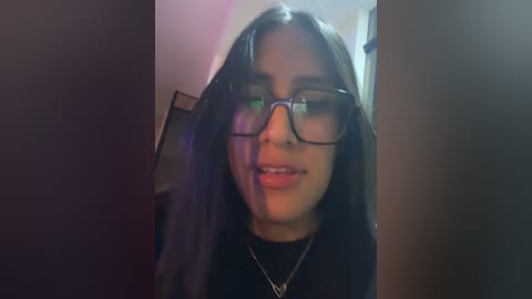 A video of a young woman with long, dark hair, wearing black-rimmed glasses and a black top, standing in a dimly lit room with blurry background.
