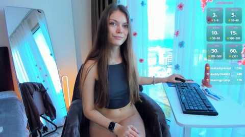 Video of a young woman with long brown hair, wearing a black crop top and beige shorts, sitting at a desk with a computer monitor displaying digital clocks.
