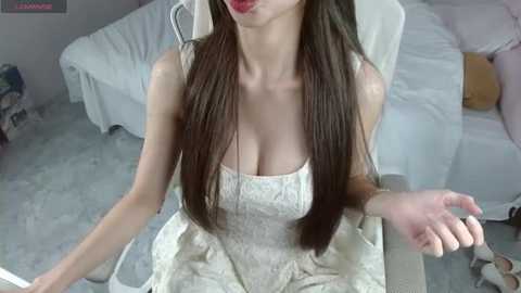 Video of a slender East Asian woman with long, straight brown hair, wearing a white, low-cut lace dress, seated on a white chair in a modern, light-colored room with a bed and toys in the background.