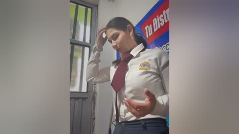 Video of a young woman in a white shirt, red tie, and navy pants, standing in a room with a window and \"Dish\" poster. She is adjusting her glasses and gesturing with her hands.