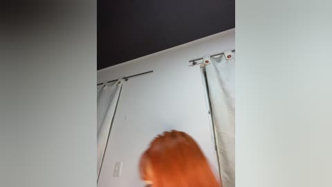 A video captures a person with long, reddish-brown hair from behind, standing in front of a white wall with a black ceiling and light gray curtains.