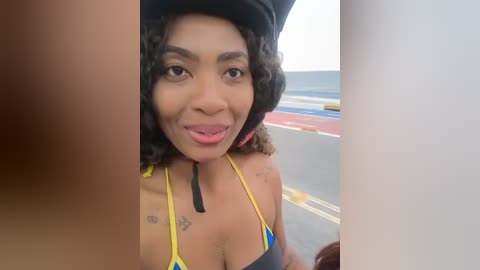 Video of a smiling Black woman with curly hair in a yellow bikini, wearing a black hat, standing on a boat deck.