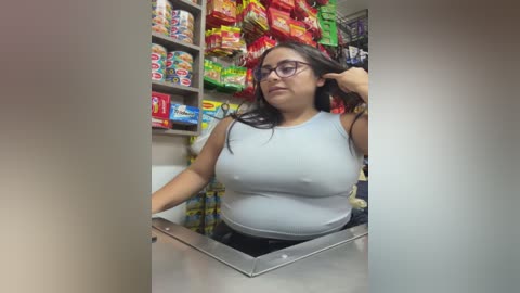Video of a Latina woman with medium skin tone, long dark hair, and glasses, wearing a tight, light blue tank top revealing her large breasts, standing in a colorful, cluttered convenience store aisle filled with snacks.