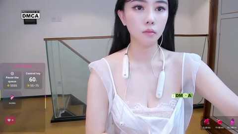 Video of a young East Asian woman with long black hair, wearing a sheer white dress, white earbuds, and a white phone. She stands in a modern, well-lit room with a glass staircase.