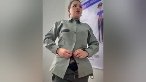 Video of a Latina woman in a gray military-style uniform with epaulettes, adjusting her shirt buttons in a professional setting, with a poster of a woman in military gear in the background.