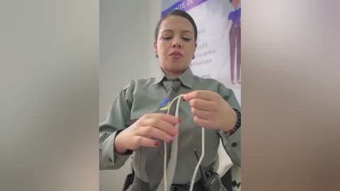 Video of a woman in a grey uniform with a black tie, holding a white cable, standing in a white-walled room with a poster.