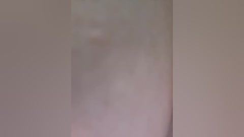 A blurred video featuring a person standing in a dimly lit room with beige walls and a corner on the right. The individual is indistinct, with a soft, diffused appearance.