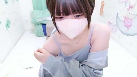 Video of an Asian woman with a short bob haircut, light skin, and wearing a gray off-shoulder top and white mask, standing in a tiled bathroom with floral wallpaper.