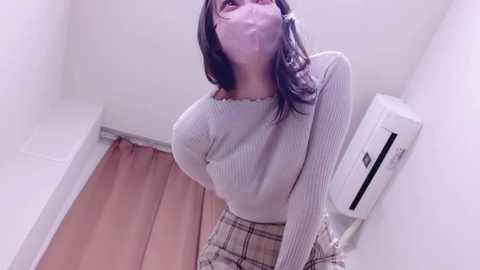 Video of a young woman with long, dark hair, wearing a gray ribbed sweater and plaid skirt, standing in a small, white-walled room with a wooden floor and a white air conditioner.