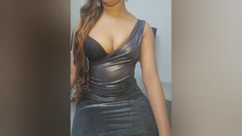 Video of a woman with long, wavy brown hair wearing a tight, shiny gray dress that reveals a black bra, sitting against a plain beige wall.