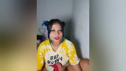 Video of a smiling, medium-skinned woman with blue hair in twin buns, wearing a yellow tie-dye \"Bad Bunny\" shirt, in a dimly lit room.