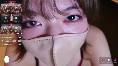 A close-up video of an Asian woman with short brown hair, wearing a beige mask, tear-stained face, and intense blue eyes.