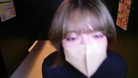 Media: Video of a person with short, straight blonde hair, wearing a black shirt, with a white cloth covering their nose and mouth, standing indoors in a dimly lit room with posters on the wall.