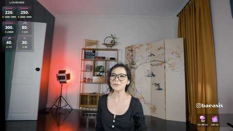 Video of a middle-aged Asian woman with glasses, wearing a black top, in a modern, minimalist room with a light-colored floor, a shelf, and a folding screen.