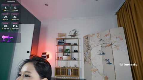 Video of an Asian woman with black hair, in a modern living room with a wall-mounted TV displaying a live view, and a large, abstract art screen.