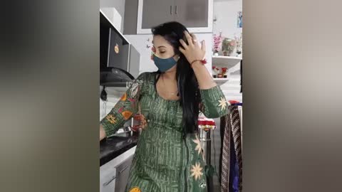 Video of a South Asian woman with long black hair, wearing a green and yellow patterned sari, mask, and headscarf, adjusting her hair in a modern kitchen.