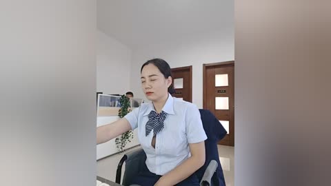 Video of an Asian woman with fair skin, dark hair, wearing a white blouse and navy tie, seated in an office chair, in a modern, minimalist room with wooden doors and green plants.
