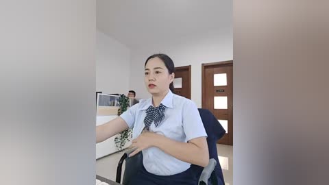 Video of a middle-aged Asian woman in a light blue blouse, seated in a wheelchair, in a modern, minimalist office with white walls, wooden door, and green potted plant.