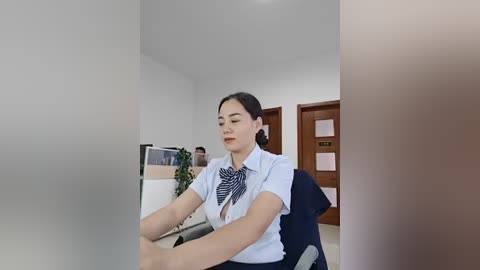 Video of a young East Asian woman with fair skin, dark hair in a bun, wearing a white blouse with a blue tie, seated in an office chair, with a blurred, beige wall and wooden door in the background.