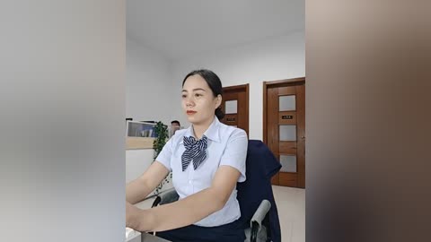 Video of an East Asian woman with fair skin and straight black hair, wearing a white blouse with a black patterned bow tie, seated in an office chair. Background includes a wooden door and white walls.