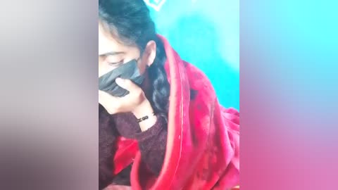 Video of a young South Asian woman with dark hair, wearing a red shawl, black mask, and dark top, leaning against a turquoise wall, looking distressed.
