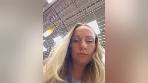 Video of a blonde woman with long hair, wearing a light blue shirt, looking serious inside a warehouse with industrial lighting and exposed ceiling beams.