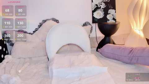 Video of a cozy bedroom with a white moon-shaped cushion, black nightstand, white and purple pillows, and floral artwork.