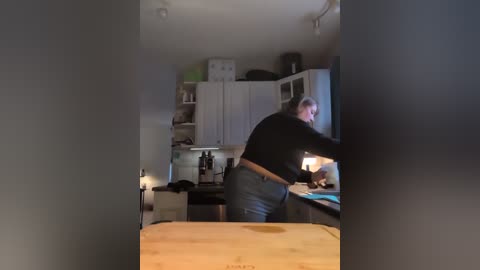 Video of a man with glasses, short gray hair, wearing a black sweater and jeans, preparing food at a wooden kitchen counter. Background shows a white cabinet, bottles, and a green bag.