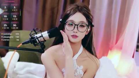 Video of a young Asian woman with long brown hair and glasses, wearing a white lace top, sitting in front of a microphone, smiling, in a cozy, dimly-lit room.