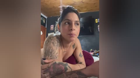 A video of a tattooed woman with dark hair in a messy bun, wearing a burgundy top, resting on a bed with a wooden ceiling. The room has dark walls, a TV, and various small items.