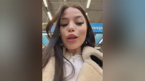 Video of a young woman with long brown hair, wearing a beige fur coat, white top, and necklace, posing indoors with a blurred background.