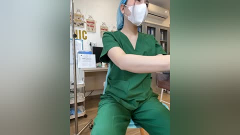 Media: Video of a female nurse in green scrubs, face mask, and blue cap, seated at a desk in a medical office. Background includes office supplies, signs, and a window.