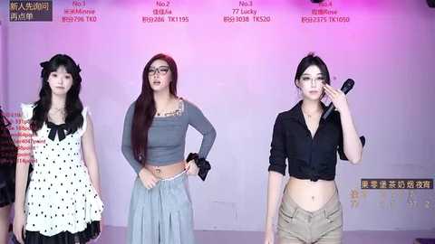 Video of three young Asian women in casual attire, standing against a white wall with red text. One wears a polka-dot dress, another a grey crop top and skirt, and the third a black shirt and shorts.