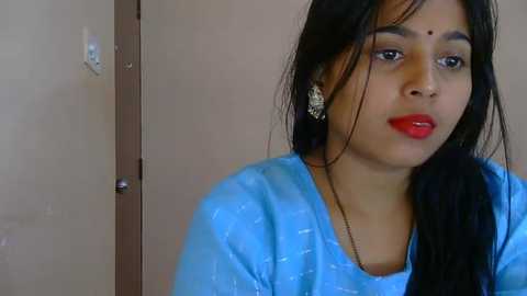 Video of a young South Asian woman with long black hair, wearing a blue saree, red bindi, and large silver earrings. She stands in front of a beige wall with a door in the background.