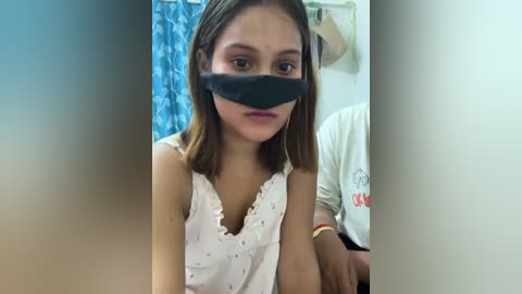 Video of a young Southeast Asian woman with light brown skin, wearing a white sleeveless blouse, black face mask, and brown hair. Background features a blue curtain and white wall.