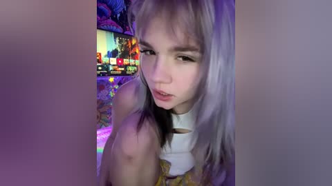 Video of a young woman with long, platinum blonde hair, fair skin, and a slender build, wearing a white top, sitting in a dimly lit room with colorful, neon-lit gaming setups in the background.