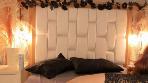 Video of a modern, cozy bedroom with a white tufted headboard, black pillows, and warm lighting. Ornate floral arrangements flank the bed, enhancing the intimate atmosphere.
